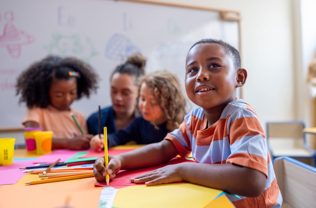 Children sitting with a teacher with a child in the foreground writing on a piece of paper